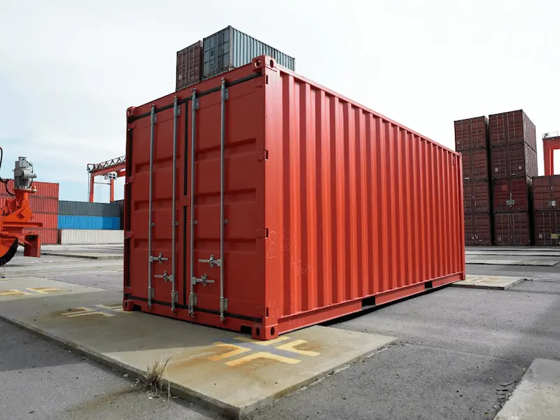 Clean, red 20ft shipping container sitting alone on concrete pads in a port yard, symbolizing a new asset purchase.