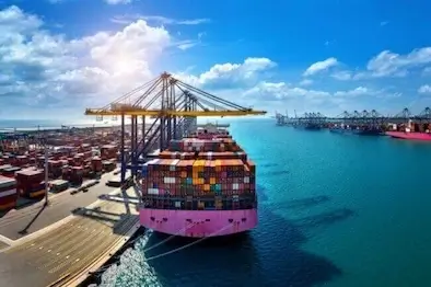 A dramatic wide-angle shot of a huge pink container ship docked at a sunny terminal, fully loaded with multi-colored containers and serviced by large gantry cranes.