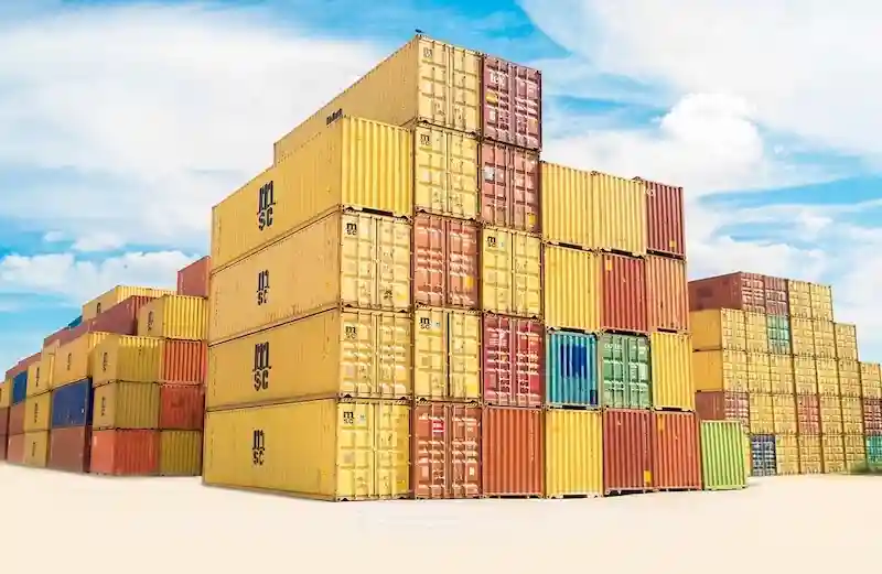 Towering stacks of multi-colored shipping containers (yellow, red, blue) in a port yard under a bright, sunny sky.