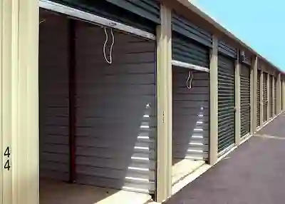 Long row of traditional metal self-storage units with dark green roll-up doors, showing open and closed bays.