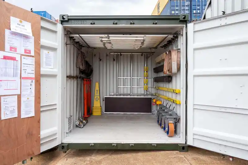 Highly organized, modified container interior with overhead shelving, wall racks, safety signs, and various lifting equipment stored.