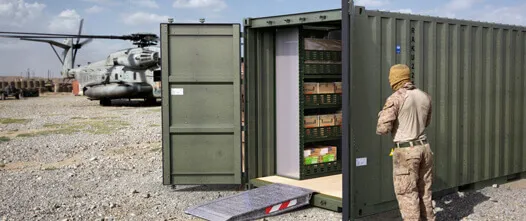 Olive drab container open in a desert field with shelves of supplies; a soldier and large military helicopter are nearby.