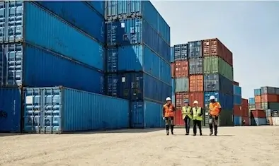 Four employees in hard hats and safety vests walking in front of towering stacks of blue and red shipping containers in a sunny cargo yard.