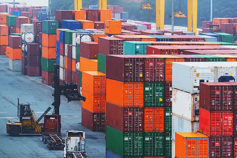 Busy cargo port with hundreds of stacked containers in various colors, forklifts, and trucks, showing high market activity.