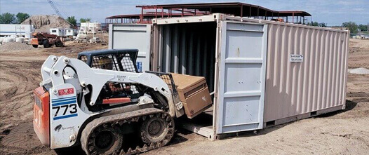 Skid-steer loading large equipment into an open tan shipping container on a dirt construction or agricultural site.