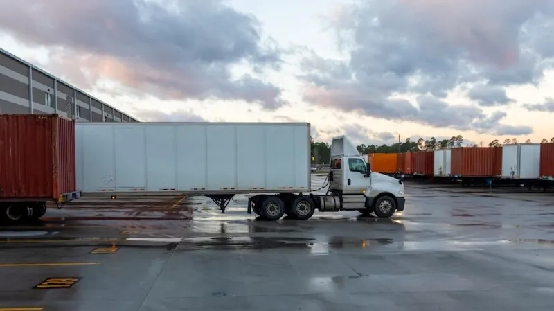 White semi-truck pulling a trailer in a wet depot yard at dusk, surrounded by stacked shipping containers.