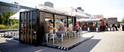 Black shipping container restaurant with a fold-down patio deck, awning, and customers seated at a bar in a sunny urban setting.