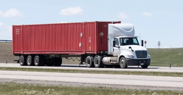 A white semi-truck pulling a flatbed trailer loaded with a long, red 40ft high cube shipping container on a highway through grassy terrain.