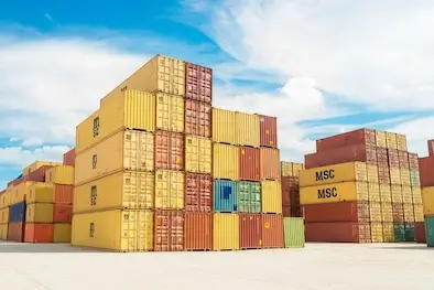 Wide, sunny view of towering stacks of multi-colored shipping containers (yellow, red, blue) in a major intermodal port, symbolizing the scale of global trade.