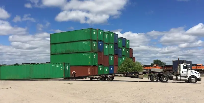 White semi-truck using a tilt-bed trailer to drop a large green shipping container on a dirt lot near stacked inventory.