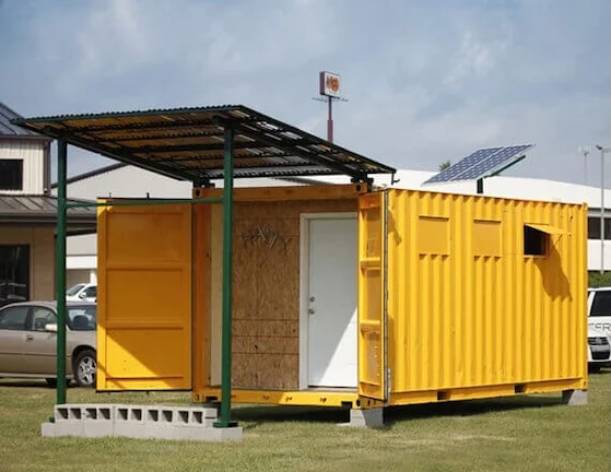 Yellow modified shipping container with a covered porch and solar panel, used as an emergency shelter or field clinic.