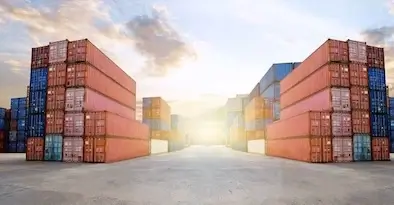 A dramatic, wide-angle view of a cargo port at sunrise/sunset, showing towering stacks of red, blue, and white shipping containers under a cloudy, bright sky.