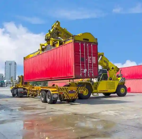 Yellow reach stacker lifting a red shipping container onto a chassis trailer in a port yard, symbolizing loading logistics.