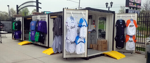 Two white shipping containers opened to create a retail shop, displaying merchandise racks at an event or outdoor venue.