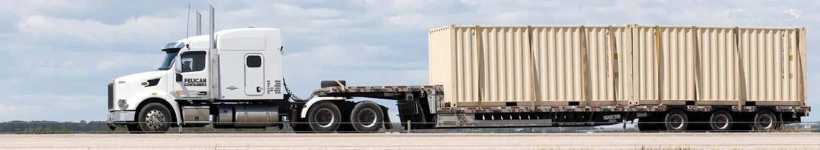 A white Pelican Containers semi-truck hauling two stacked beige 20ft storage containers on a flatbed trailer down a highway under a cloudy sky.