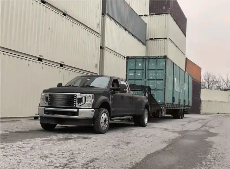 Dual-wheeled pickup truck towing a green 20ft shipping container out of a depot yard with stacked beige containers.