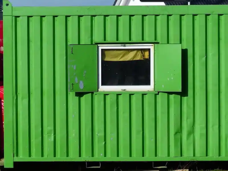 Close-up of a green shipping container with a white window frame and two metal protective shutters opened.