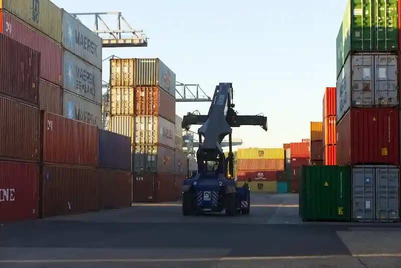 Large blue reach stacker machine operating between towering stacks of multi-colored shipping containers in a depot.