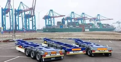 Blue container chassis parked in the foreground, with massive red and blue port cranes and a container ship in the background, symbolizing intermodal transport scale.