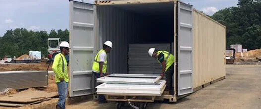 Construction workers in vests loading doors and building materials into a large 20ft shipping container on a job site.