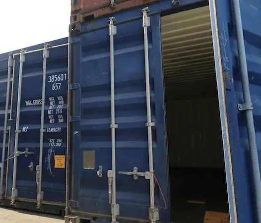 A close-up of a blue used shipping container with one door open, showing the dark interior, door locking rods, and stenciled weight specifications.