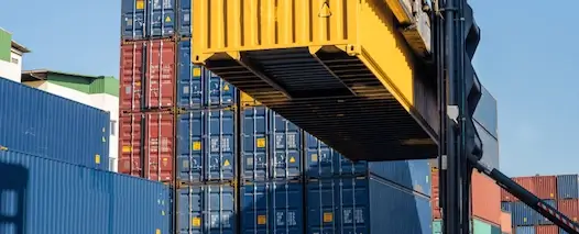 A powerful forklift (reach stacker) lifting a bright yellow shipping container over stacks of blue and red containers in a busy, sunny cargo port.