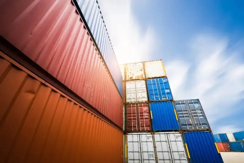 Dramatic low-angle view of multi-colored shipping containers stacked high under a bright sky, symbolizing immense mass and weight.